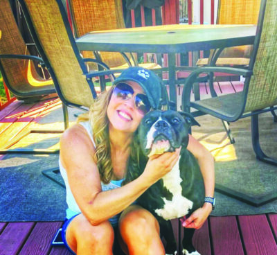 woman in tank top and shorts and sunglasses, sitting on porch hugging medium sized dog
