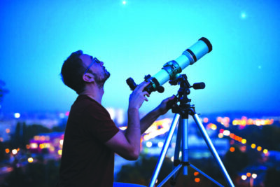 Astronomer with a telescope watching at the stars and Moon with blurred city lights in the background.