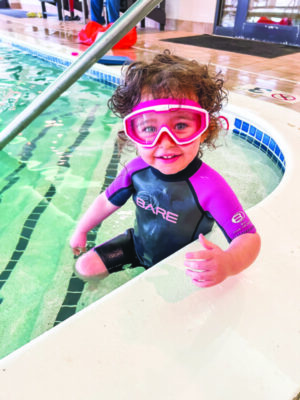 toddler wearing wet suit and goggles, sitting on steps to indoor swimming pool