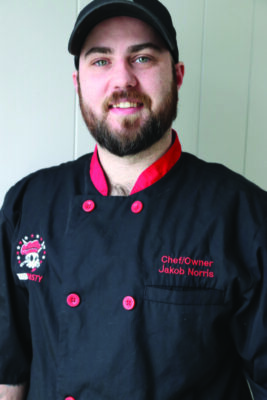 man with beard wearing black and red chef's jacket with baseball cap, slight smile to camera