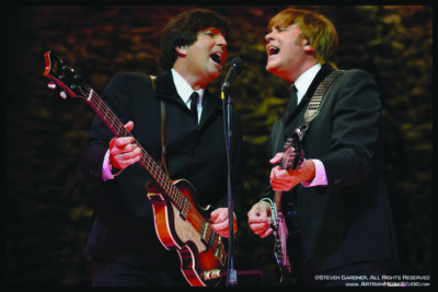 2 men playing guitar and singing into microphone, dressed in vintage suits to look like Beatles band members