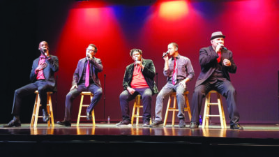 5 men sitting on wooden stools on stage, holding microphones, dressed in button up shirts and ties