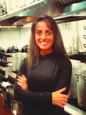 long haired woman wearing black long sleeve shirt, standing with arms crossed in kitchen, smiling