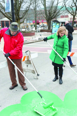 a man and a woman wearing winter coats and gloves, holding long paint rollers to paint green shamrock on pavement
