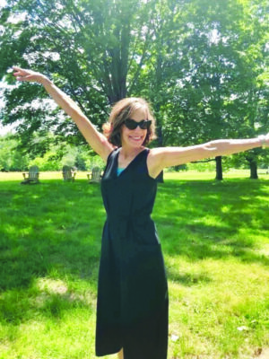 woman wearing sunglasses and black summer dress, standing outside with arms outstretched, smiling