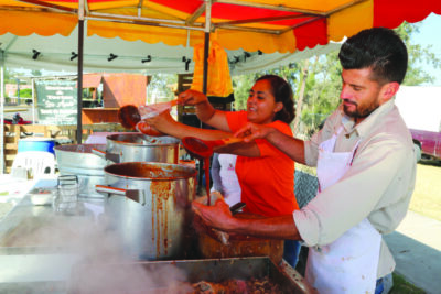 man and a woman under a red and yellow event tent, serving birria out of large pots