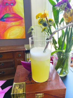 yellow cocktail in tall glass with straw, sitting on table beside jar of flowers