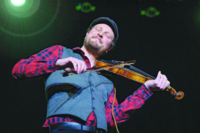 bearded man wearing plaid shirt, vest and cloth cap, playing violin under stage lights