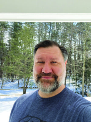 headshot of man with beard, wearing t-shirt, standing in front of window showing snowy woods outdoors