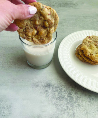 hand dipping cookie into glass of milk beside plate containing cookies