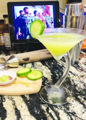cocktail in martini glass with cucumber garnish, on counter cluttered with ingredients