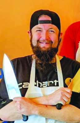 man holding knives with arms crossed, wearing apron and backwards baseball cap, smiling