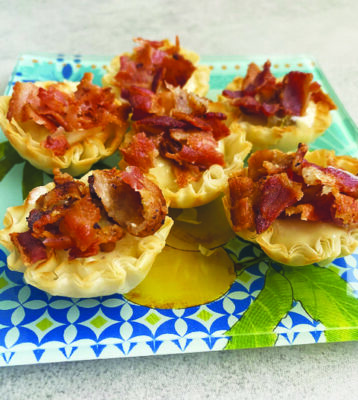 6 cheese tarts with crimped edges, topped with bacon, arranged on colorful square plate