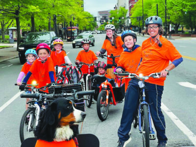 2 woman, 6 children, 1 dog, stopped on bikes in bike lane on city street, all wearing orange shirts and helmets