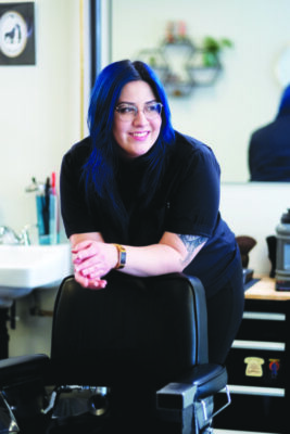 young woman with glasses, blue tinted hair, leaning on back of hairdresser chair in barber shop
