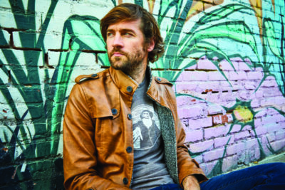 man wearing t shirt and leather jacket, has beard, sitting against wall mural, looking to the side