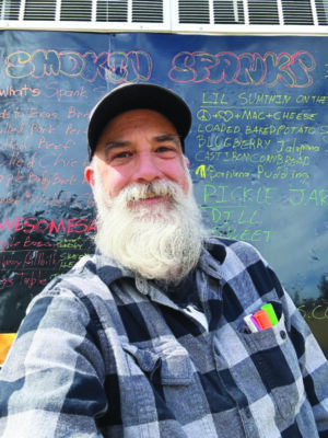 man with bushy white beard, wearing baseball cap, sitting in front of blackboard with menu written in colored chalk