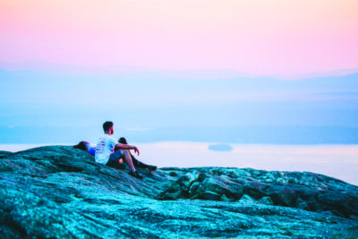 man and woman sitting on rocks at mountain summit with glow of sunrise behind them