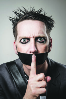 man with dark eyeliner, tape across mouth, holding finger up to mouth