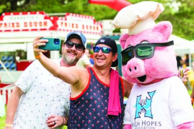 two men taking selfie with costumed mascot, pig wearing chef's hat and sunglasses