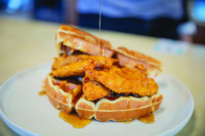 plate with large belgian waffles topped with crispy chicken, syrup being poured on top