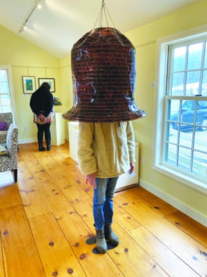 large oblong shaped hat hanging from ceiling, woman standing under had covered down to shoulders