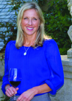 woman in blue blouse, standing outside near stone wall, foliage in background, holding large wine glass, smiling