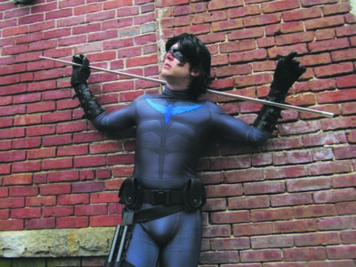 man dressed at comic character Nightwing in spandex suit and black eye mask, leaning against brick wall with pole