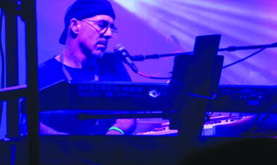 man sitting at keyboard singing into microphone under colored lights