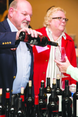 man standing behind table full of wine bottles, pouring wine into someone's cup, woman standing beside him