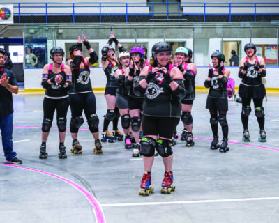 roller derby team standing in line and cheering for team mate standing in front with her hands positioned to form a heart shape