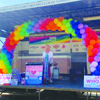 person standing on stage with Nashua Pride banner behind and colored balloons making rainbow across stage