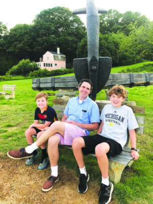 man and two boys sitting on base of anchor monument on lawn outside of building