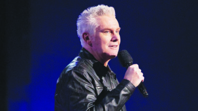 comedian Brian Regan in leather coat, holding microphone