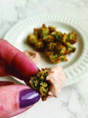 close up of thumb and forefinger holding piece of breaded and baked okra dipped in sauce