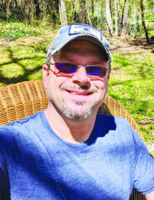 headshot of man with short beard, going a little gray, sitting in wicker chair outside on sunny day wearing sunglasses and baseball cap