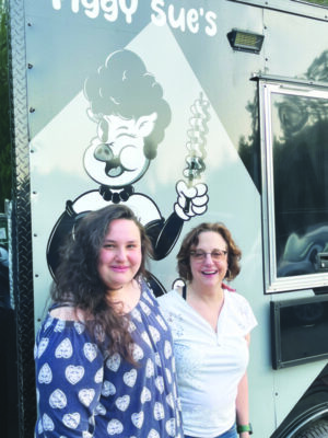 2 women standing in front of food truck painted with cartoon female pig wearing dress and holding up bacon