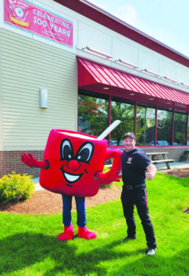 man standing outside building beside red arrow mascot of red mug with smiling face painted on
