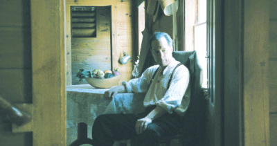 Man dressed in white shirt, suspenders, dark pants, sitting in shadowed room with wooden walls, beside table with wooden bowl of vegetables, old time feel