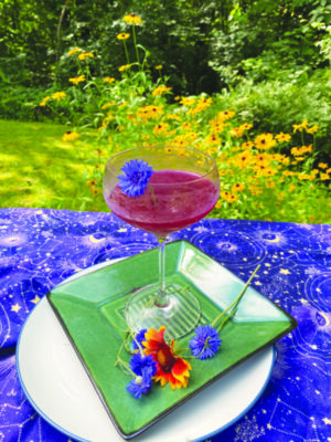 wine glass half filled with dark purple margarita, garnished with small blue flower, glass sitting on plate on table outside grass and black-eyed susan flowers in background
