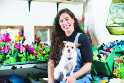 Woman sitting in art studio with colorful materials on table behind her, dog on her lap