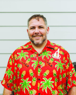 headshot of smiling middle aged man wearing bright patterned Hawaiian style shirt with 603 Brewery logo printed on left chest, outside in front of paneled wall