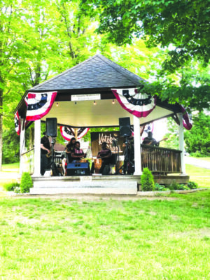 band members with instruments in gazebo decorated with lights and red, white, and blue banners, grassy lawn and trees behind