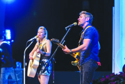 male and female performers playing guitars at separate microphones, on stage at night
