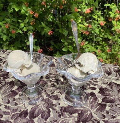 2 fancy stemmed bowls, each containing 2 scoops of light colored ice cream and spoons, sitting on embroidered table cloth outside
