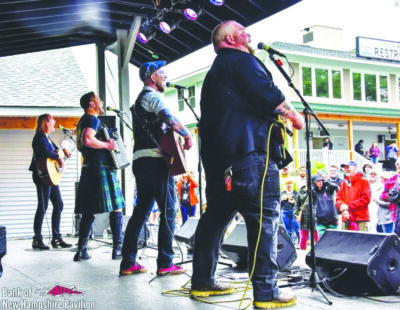 four band members on covered outdoor stage, playing in front of crowd in town, seen from back