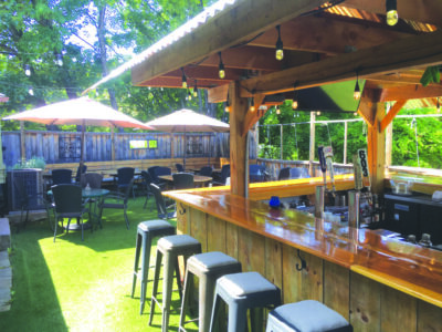 restaurant outdoor patio with wooden bar under awning, tables under large umbrellas, enclosed by fence, trees beyond
