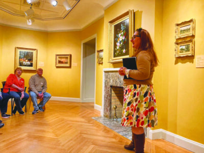 standing woman talking to people lined up in chairs in art gallery, small and medium sized paintings in ornate gold frames along the walls