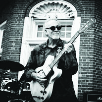 older man standing outside of brick building playing guitar, wearing sunglasses