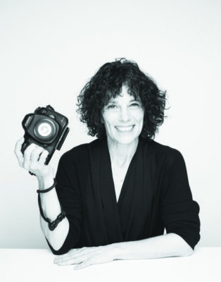 black and white portrait of women with one hand on table, holding up camera in right hand, smiling, blank background